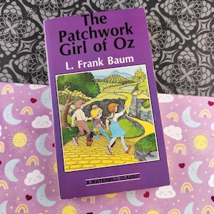 Może przedstawiać: Fioletowa książka w twardej oprawie zatytułowana "The Patchwork Girl of Oz" autorstwa L. Franka Bauma. Okładka przedstawia ilustrację dzieci na żółtej drodze z cegieł. Książka zawiera napis "A Watermill Classic".