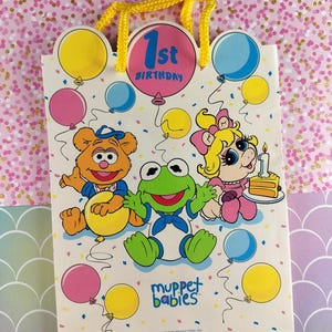 Puede incluir: Una bolsa de regalo vintage de los Muppet Babies para el 1er cumpleaños. La bolsa presenta ilustraciones de Fozzie Bear, Kermit la Rana y Miss Piggy, junto con globos y confeti. La bolsa tiene un asa de cuerda amarilla.
