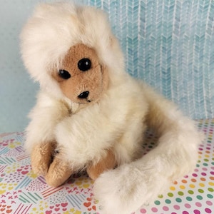 Pode incluir: Um macaco de pelúcia creme com rosto, mãos e pés castanhos claros. O macaco tem olhos pretos e uma cauda longa e fofa. O brinquedo está sentado em uma superfície estampada colorida.