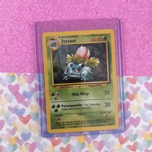 May include: A Pokémon trading card featuring Ivysaur, a green and blue creature with a bulb on its back. The card is in a protective sleeve. The card text includes "Vine Whip" and "Poisonpowder". The background is pink with floral patterns.