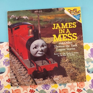 Puede incluir: Portada de libro infantil con un tren de juguete rojo y el título "James in a Mess" en letras amarillas. También se ve el texto "and Other Thomas the Tank Engine Stories". El libro está basado en la serie Railway.