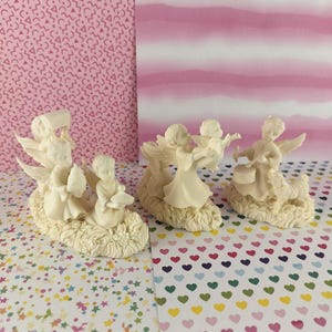 May include: Ivory-colored angel figurines in various poses, some holding objects, others with wings spread. The figurines are set on a textured base, with a soft, creamy hue. The background features patterned paper.