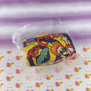May include: A sealed plastic bag containing a colorful, rectangular toy. The toy features a cartoon image of a child with sunglasses and the words "Too Cool For School" in red and blue on a yellow background. The toy is a Wendy's Kids Meal item.