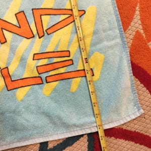May include: A light blue beach towel with the word "WAVE" in orange letters outlined in black, with yellow and pink accents. The towel measures approximately 58 inches in length.