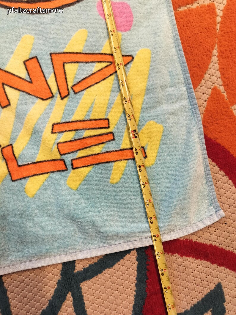 May include: A light blue beach towel with the word "WAVE" in orange letters outlined in black, with yellow and pink accents. The towel measures approximately 58 inches in length.