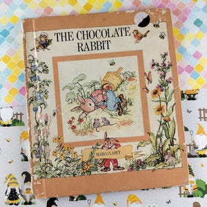 Op de afbeelding: Vintage kinderboek getiteld "The Chocolate Rabbit" door Maria Claret. De omslag toont een grillige illustratie van een konijn, een kind en andere dieren in een tuin. Het boek heeft een bruinachtige kaft met slijtage.