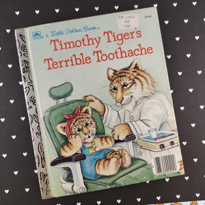 May include: A Little Golden Book titled "Timothy Tiger's Terrible Toothache." The cover features an illustration of a tiger cub in a dentist's chair, with a concerned adult tiger examining its teeth. The book has a light blue cover.