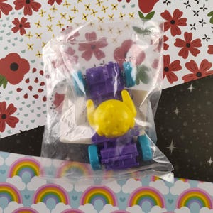 May include: A purple toy car with yellow wheels and a yellow character in the driver's seat. The car is in a clear plastic bag.