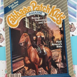 Puede incluir: Una portada de la revista Cabbage Patch Kids con una muñeca montando un caballo en una calle adoquinada de Boston. El título de la revista es "Cabbage Patch Kids" con el subtítulo "Boston - Ayer y Hoy". El texto "Paul Revere's Ride" también es visible en la portada.