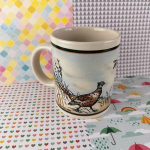 May include: Cream-colored ceramic mug with a pheasant illustration. The mug features a detailed painting of a pheasant in a field with tall grass and a blue sky. The mug has a green and black trim.