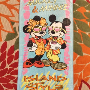 May include: A light blue beach towel featuring Mickey and Minnie Mouse in sunglasses. The towel has the words "Mickey & Minnie" and "Island Style" in a colorful design. The cartoon characters are wearing tropical-themed outfits.