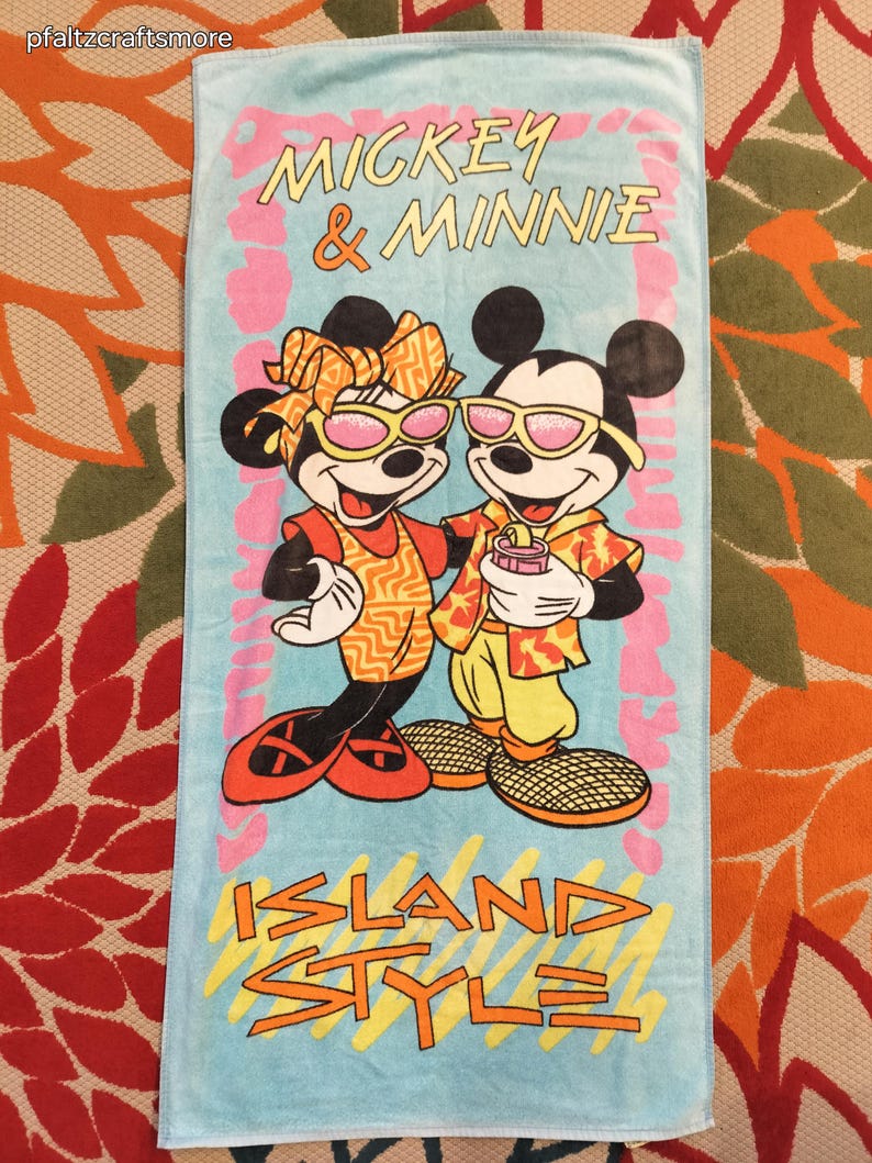 May include: A light blue beach towel featuring Mickey and Minnie Mouse in sunglasses. The towel has the words "Mickey & Minnie" and "Island Style" in a colorful design. The cartoon characters are wearing tropical-themed outfits.