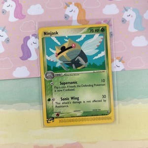 Pode incluir: Um cartão de troca Pokémon Ninjask com uma borda amarela, apresentando uma criatura semelhante a um inseto com asas. O cartão exibe o texto "Ninjask" e "70 HP", juntamente com descrições de ataque. O cartão está em uma manga protetora.