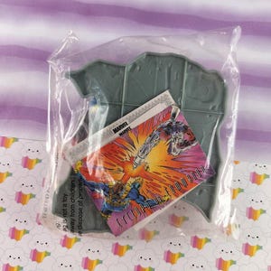 May include: A Marvel Comics collectible, sealed in a clear plastic bag. The item includes a gray plastic base and a trading card featuring Cyclops and another character in a battle scene. The card has the Marvel logo.