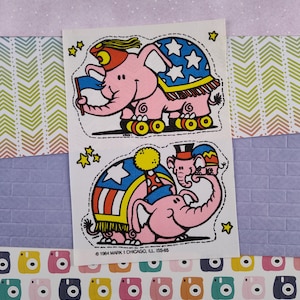 May include: Two pink elephant stickers with patriotic designs. One elephant is on roller skates and holding a flag. The other elephant is wearing a hat and a cape with stars. The text "©1984 MARK 1 CHICAGO, ILL. ISS-65" is printed on the sticker.