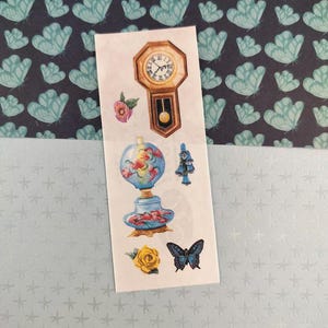 Peut inclure: Une planche d'autocollants avec diverses illustrations de style vintage. Les motifs comprennent une horloge, une lampe en forme de globe, un papillon bleu, une rose jaune et d'autres éléments floraux. Le fond est bleu clair avec des motifs d'étoiles.