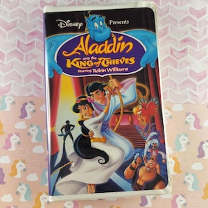 Puede incluir: Una cinta VHS de Disney de "Aladdin y el Rey de los Ladrones" protagonizada por Robin Williams. La portada presenta a Aladdin y Jasmine, con el Genio y otros personajes. El fondo es un patrón rosa y blanco.