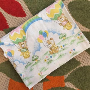 May include: White baby sheet with a whimsical design featuring teddy bears, balloons, and a rainbow. The bears are depicted in hot air balloons and holding colorful balloons. The sheet has a light blue sky and green grass.
