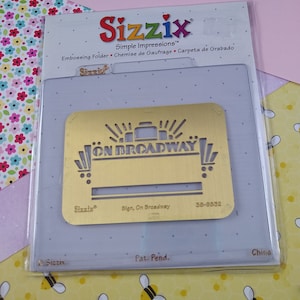 May include: A gold-colored metal embossing folder with the text "ON BROADWAY" in a stylized font. The folder is packaged in a clear plastic sleeve with the brand name "Sizzix" and the product name "Sign, On Broadway" printed on it.