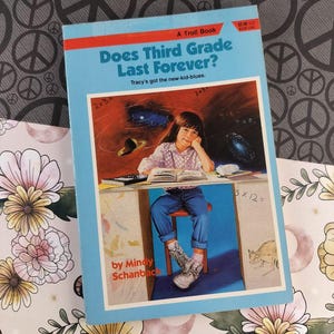 Może przedstawiać: Książka dla dzieci zatytułowana "Does Third Grade Last Forever?" przedstawia młodą dziewczynę przy biurku. Okładka jest jasnoniebieska z czerwonym i białym tekstem. Książka autorstwa Mindy Schanbac, wydana przez A Troll Book.