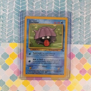 Puede incluir: Una carta coleccionable de Pokémon con Shellder, una criatura bivalva púrpura con una lengua roja, en una carta azul y amarilla. La carta está en una funda protectora. El texto de la carta incluye "Supersonic" y "Hide in Shell".