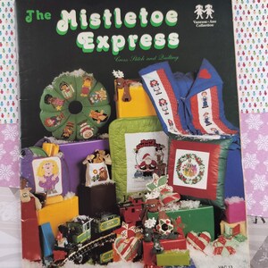 May include: A book titled "The Mistletoe Express" with a colorful cover featuring cross stitch and quilting patterns for Christmas ornaments, pillows, and a train. The Vanessa-Ann Collection.
