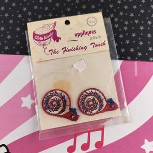 Puede incluir: Juego de apliques vintage "Cloth World" con dos diseños de caracoles. Cada caracol es rojo con un patrón en espiral azul y blanco. El embalaje incluye el texto "The Finishing Touch".