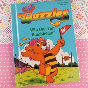 Könnte beinhalten: Ein Vintage-Buch "Wuzzles", "Win One For Bumblelion", zeigt einen Cartoon-Löwen-Bienen-Hybriden, der eine Flagge hält. Das Cover ist hellblau mit farbenfrohem Text und Illustrationen. Das Buch gehört zur Collector-Serie, Buch Nr. 4.