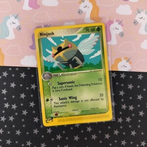 Pode incluir: Um cartão colecionável Pokémon Ninjask com uma borda amarela. O cartão apresenta uma ilustração de uma criatura semelhante a um inseto com asas. O cartão tem texto que inclui "Supersonic" e "Sonic Wing".