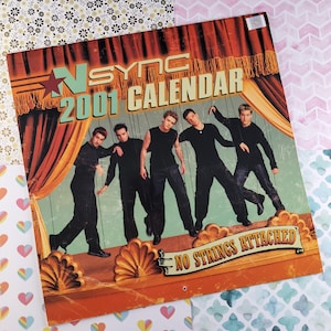 May include: A vintage 2001 calendar featuring the boy band *NSYNC. The calendar has a red curtain backdrop and the band members are dressed in black and posed as puppets with strings attached. The text "NO STRINGS ATTACHED" is printed on the bottom of the calendar.