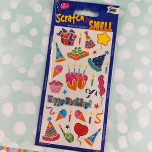 May include: A package of "Scratch N' Smell" acid-free stickers with birthday-themed designs. The stickers feature images of gifts, birthday cakes, party hats, ice cream cones, and the words "Happy Birthday!" in a colorful arrangement.