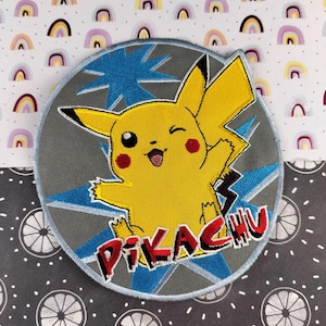 May include: Embroidered patch featuring Pikachu, a yellow creature with red cheeks, winking and smiling. The patch is circular, with a gray background and blue starburst design. The word "PIKACHU" is embroidered in red.