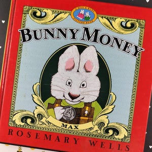 May include: A hardcover children's picture book titled Bunny Money by Rosemary Wells. The red cover features a central illustration of a white rabbit named Max wearing green overalls and holding a coin, framed by gold scrollwork and blue patterns.