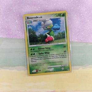 May include: A Pokemon trading card featuring Roserade lv.33. The card has a green and yellow border with an illustration of a green and white creature. The card text includes "Poison Sting" and "Spike Whip".