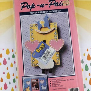 May include: A pink Pop-n-Pai craft kit for a single switch plate/frame, featuring a baby theme. The kit includes pre-stamped cut wood, instructions, diagrams, and a bonus project. The design includes a rocking horse, hearts, and a sheep.