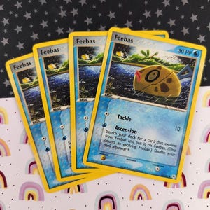 Könnte beinhalten: Vier Feebas Pokémon-Sammelkarten mit gelben Rändern sind aufgefächert. Die Karten zeigen eine Cartoon-Fisch-Illustration und Text, darunter "Tackle" und "Ascension". Der Hintergrund ist ein Sternenmuster über einem Regenbogen-Design.