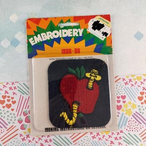 Könnte beinhalten: Ein Bügelbild-Stickerei-Patch mit einem roten Apfel und einem gelben Wurm. Der Patch befindet sich auf einem dunkelblauen Hintergrund und ist mit "EMBROIDERY IRON-ON" beschriftet. Die Verpackung enthält einen JCPenney-Aufkleber.