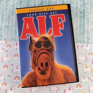 Puede incluir: Set de DVD de la primera temporada de la serie de televisión ALF. La portada presenta un primer plano de la cara del personaje alienígena sobre un fondo azul estrellado. El título "ALF" está en letras rojas grandes.