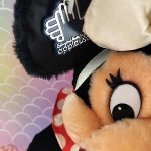 May include: Close-up of a plush Minnie Mouse doll. The doll has a black head with a white bow, a peach-colored face, and a red and white polka-dot dress. A tag with the text "Everyone loves to applause" is attached to the doll.