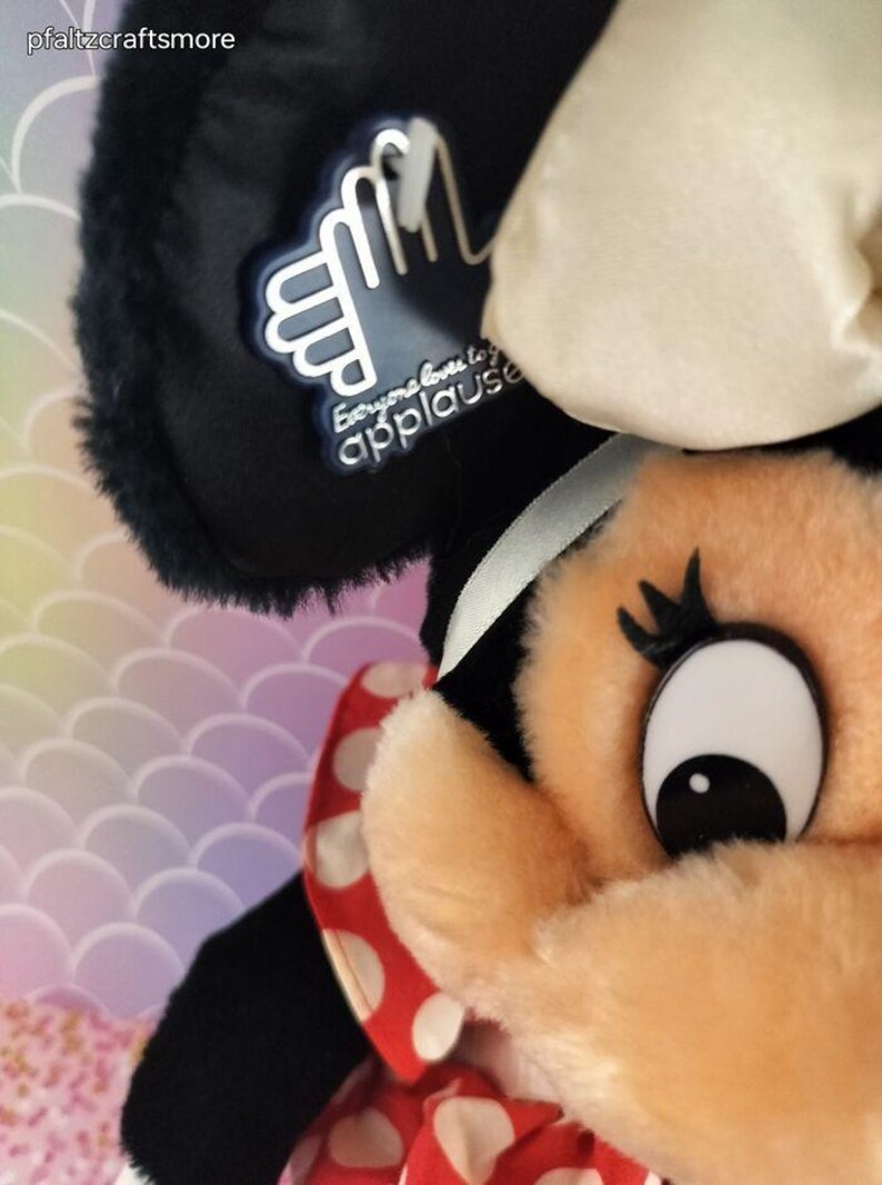 May include: Close-up of a plush Minnie Mouse doll. The doll has a black head with a white bow, a peach-colored face, and a red and white polka-dot dress. A tag with the text "Everyone loves to applause" is attached to the doll.