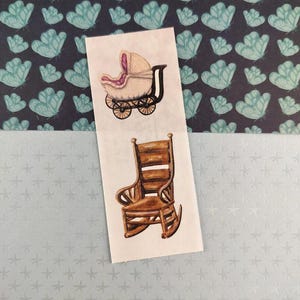 Peut inclure: Illustrations à l'aquarelle d'une poussette et d'un fauteuil à bascule. La poussette est crème avec un intérieur rose et des roues noires. Le fauteuil à bascule est marron. Le fond présente un motif de papillons et d'étoiles bleus.