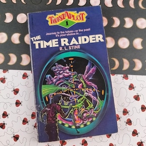 May include: A vintage paperback book titled "The Time Raider" by R.L. Stine. The cover is predominantly blue with a colorful illustration of a futuristic scene. The book is part of the "Twist-A-Plot" series.