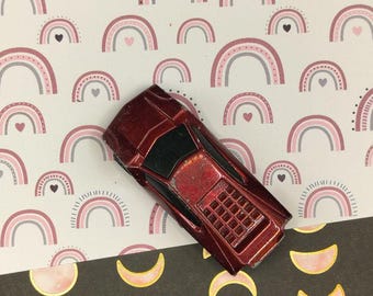 May include: A red toy car with a black windshield sits on top of a sheet of paper with a pink and gray rainbow pattern. The paper is on top of a black sheet of paper with a pink and gold moon pattern.