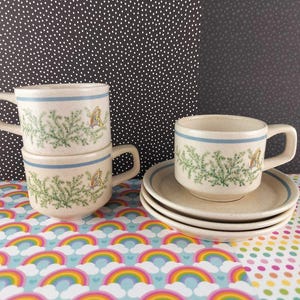 Op de afbeelding: Drie keramische kopjes en twee schoteltjes met een gespikkelde crèmekleurige afwerking. Elk stuk heeft een blauwe horizontale band bij de rand en een groen varenachtig botanisch patroon met een kleine gele vlinder. De tekst pfaltzcraftsmore is zichtbaar.