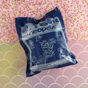 Puede incluir: Un paquete de juguetes Neopets azul oscuro con varias ilustraciones de personajes de dibujos animados. El paquete incluye el texto "Neopets" y "Código de premio virtual incluido". El fondo presenta una superficie rosa con purpurina dorada y un patrón de vieira pastel.