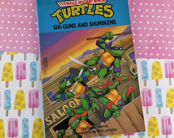 May include: A vintage paperback book titled "Teenage Mutant Ninja Turtles: Six-Guns and Shurikens." The cover features the four turtles in action, with a saloon in the background. The book is set against a pink and popsicle-patterned backdrop.
