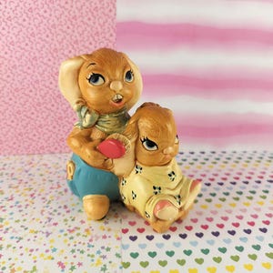May include: A ceramic figurine set featuring two brown bunnies. One bunny is wearing blue overalls and holding a red heart. The other bunny is wearing a yellow dress with blue floral accents. The bunnies have blue eyes and are on a patterned surface.