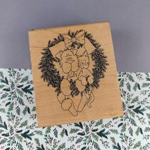 May include: A wooden stamp featuring a dog and cat inside a wreath with a bow. The wreath is made of evergreen branches. The stamp is on a patterned fabric with green and red floral designs.