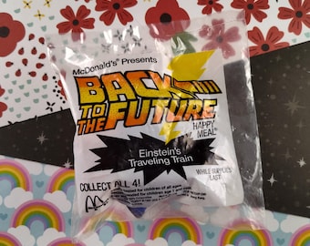 May include: A plastic bag containing a McDonald's Happy Meal toy from the Back to the Future movie. The toy is a yellow and black train with the words "Einstein's Traveling Train" printed on it. The bag also has the words "McDonald's Presents Back to the Future Happy Meal" printed on it.