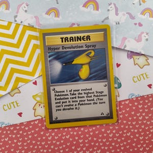 Può includere: Una carta Allenatore Pokémon gialla e nera con il testo "TRAINER" e "Hyper Devolution Spray". La carta presenta un'illustrazione di uno spruzzino giallo con un beccuccio nero.
